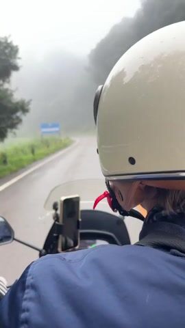 A first-person perspective video shows a person riding a motorcycle on a winding road with a white stripe down the middle. The rider is wearing a white helmet with black accents and a blue jacket. A smartphone is mounted on the handlebars in front of the rider. The road is wet and surrounded by lush green vegetation and trees, which are partially obscured by a thick fog. Yellow warning signs indicating curves in the road are visible along the roadside. The sky is overcast, and the overall atmosphere is misty and damp.
