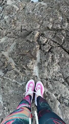 The video opens with a top-down view of a person's feet, clad in bright pink and white athletic shoes and colorful, patterned leggings, standing on a rugged, dark grey rock formation. In the background, white foamy water is visible.

As the video progresses, a large wave crashes in from the direction of the sea, engulfing the person's feet and the surrounding rocks in a burst of white foam and turbulent water. A sound akin to a surprised gasp or exclamation, "Ah!", is heard at this moment. The wave then recedes, leaving behind foamy water swirling over the textured rock surface, with the person's now-wet shoes still visible.

The scene depicts a rocky shoreline encounter with ocean waves, likely in Seoca, Montenegro. The time of day appears to be daylight.