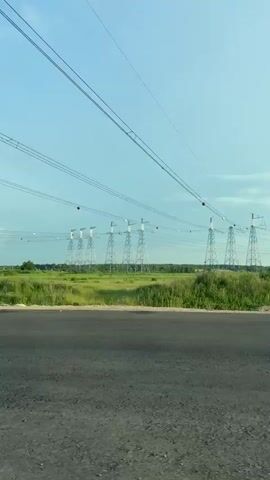 The video begins with a wide shot of a rural landscape. Power lines stretch across the frame, supported by tall metal towers in the distance. A paved road is visible in the foreground, with a grassy field and trees beyond. The sky is a clear blue with a few wispy clouds. As the video progresses, the camera appears to be inside a moving vehicle. The perspective shifts, showing the road ahead and a side mirror reflecting the passing scenery. Several power line towers are visible in the background, and the vehicle is driving on a highway with other cars in the distance. The sun is visible in the sky.