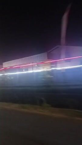 The video captures a night scene from a moving vehicle, likely a motorcycle, judging by the visible handlebar mirror and the perspective. The camera is pointed slightly to the right, showing a road in the foreground with a dark grassy area and a building with red and white lights in the background. Streaks of light suggest motion blur. As the vehicle continues, the scene shifts to reveal a wider landscape with a Ferris wheel and numerous lights, indicating a nighttime event or fairground. The sky is dark. The quality is low and grainy, with significant motion blur throughout.