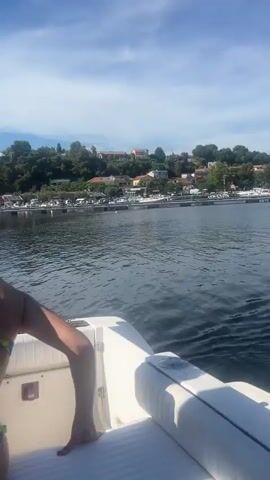 The video begins with a rear view of a woman in a bikini on a boat, looking out at the water and a town in the distance. The next clip shows three people inside the cabin of a boat, two women and a man. One woman is wearing a bikini, and the other is in swimwear. The man is at the helm of the boat. The third clip shows a man driving a boat, with a blue towel draped over a railing. The final two clips show a view of several sailboats docked in a harbor, with the water reflecting the blue sky. The video appears to be shot from a boat moving through the water.