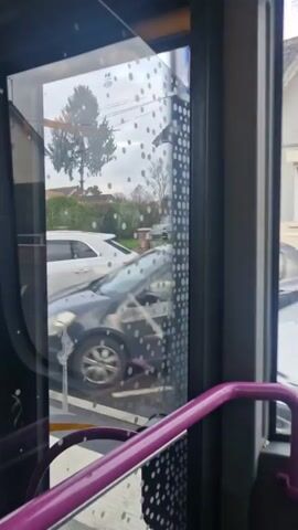 The video shows a view from inside a bus moving down a residential street. The camera faces a window covered in a pattern of small, opaque circles. Through the window, cars parked along the side of the road and houses are visible. The sky is overcast. The bus's purple handrail is visible in the foreground.