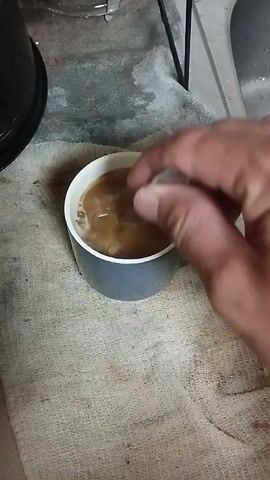 A hand is seen stirring a light brown liquid in a grey mug with a spoon. The mug is on a white towel, which is on a speckled grey surface. In the background, a black object and a silver sink are visible. The liquid appears to be coffee with milk or creamer.