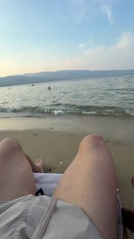 The video opens with a first-person perspective of someone relaxing on a beach chair, facing the ocean. The camera is positioned to capture the lower half of the person's legs and torso, clad in light-colored shorts. The sandy beach stretches out, with gentle waves lapping at the shore. In the background, several people are visible swimming and wading in the water. The ocean extends to the horizon, where mountains can be seen in the distance under a clear, pale blue sky with scattered clouds. The sun appears to be setting or rising, casting a soft light on the scene. The camera slowly pans to the left, revealing more of the beach and the shoreline, with beach chairs and umbrellas dotting the landscape. The focus remains on the serene beach environment, emphasizing the relaxing atmosphere.