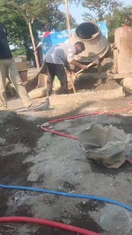 The video shows a construction site. There are two men working. One man, wearing a dark shirt and tan pants, stands looking up and to the left. The other man, wearing a light shirt and dark pants, is bent over and appears to be using a shovel to mix something in a cement mixer. A red bucket is near him. The cement mixer is a large, cylindrical, metal drum on a stand. There are bags of what appear to be cement or sand on the ground. Red and blue hoses are also visible on the ground. The environment is outdoors, with trees and a blue banner visible in the background. The ground is covered with dirt, gravel, and construction materials.