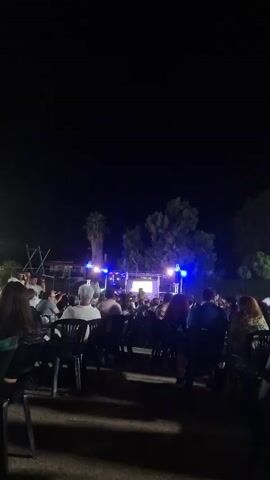 The video shows an outdoor gathering at night, with a stage set up in the background. A group of people are seated at tables, facing the stage. The stage is lit with blue lights, and there appears to be a screen or projector setup. Several palm trees and other foliage are visible behind the stage, silhouetted against the dark sky. Some individuals are standing and talking, while others are seated and watching the stage. The overall atmosphere seems to be a casual event or performance.