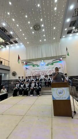 A man stands at a podium and speaks into a microphone. He is wearing a grey shirt and glasses. Behind him, a group of people in graduation gowns and caps are seated. A banner with Arabic text and images of Palestinian flags hangs behind the seated graduates. The room has a high ceiling with decorative lights.