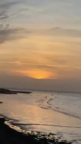 The video shows a body of water at sunset. The sky is filled with streaks of orange and grey clouds, partially obscuring the sun. The water is calm, reflecting the colors of the sky. Along the shore, there are dark rocks and sand. A line of poles extends into the water, possibly marking a path or boundary. The overall scene conveys a sense of tranquility.