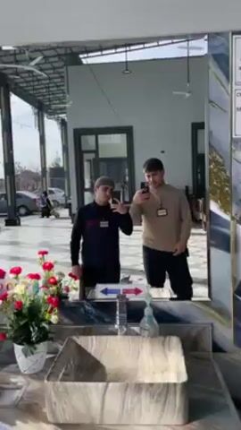Two men are taking a selfie in front of a large mirror in what appears to be a public facility, possibly a mosque or religious center, as suggested by their attire. The man on the left is wearing a dark blue jacket with red and white stripes and a taqiyah, while the man on the right is wearing a tan sweater. Each has a lanyard with an ID card around their neck.

In the background, visible through a doorway or opening, there is an outdoor area with cars parked, a few people walking, and simple landscaping. A small white van drives by.

In the foreground, below the mirror, there is a modern sink with decorative elements, a faucet, hand sanitizer, and a flower arrangement. Directional signage with arrows is displayed above the sink. The natural lighting suggests it is daytime, and the overall ambiance is clean and well-maintained.