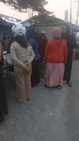A group of people are standing on a brick walkway in front of stalls or shops. The people are mostly women, some wearing hijabs. One woman is wearing a white helmet. There are tables set up, with items displayed on them, possibly for sale. One table has a white cake. A young boy is standing near a table. The background shows buildings and trees. The lighting suggests it is either early morning or late afternoon.