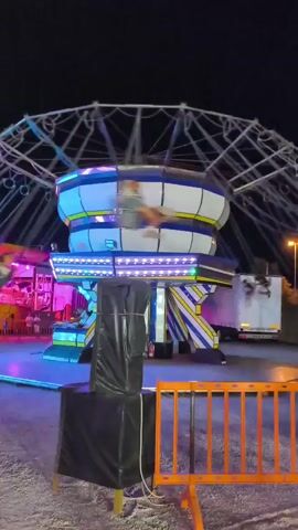 The video captures a nighttime scene at what appears to be a fair or amusement park. In the foreground, a black tarp-covered object stands on a wooden base. A bright orange barrier is visible in front of it. The central focus is a spinning amusement park ride. The ride features a central column with yellow and blue striped supports, topped by a cylindrical structure with blue and white segments and yellow trim. The structure is illuminated with bright white and purple LED lights. The ride's arms extend outwards, holding seats or gondolas that are blurred due to the motion. Several figures are visible in the blurred seats, suggesting people are riding it. In the background, a large Ferris wheel structure is visible, also illuminated. To the left, another amusement ride with pink and purple lighting is partially visible, with people standing nearby. A white truck is parked on the right side of the frame. The overall environment suggests a lively atmosphere at an outdoor nighttime event.