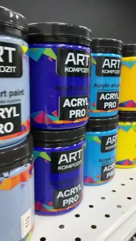 The video displays shelves filled with jars of acrylic art paint. The jars are cylindrical with black lids and feature colorful labels. The labels have the brand name "ART KOMPOZIT" and "ACRYL PRO" prominently displayed. The paint colors visible on the shelves include blue, yellow, orange, green, black, pink, and red. The jars are stacked in pairs on each shelf. The shelves are white and perforated with small holes. In the background, a gray wall is visible, and further back, a counter area with some electronic equipment and a price tag can be seen. The video pans across the shelves, showcasing the variety of paint colors. There are no people visible in the video.