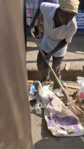 An elderly man is seen sweeping trash on a street near a market. He is wearing a white t-shirt, dark trousers, black rubber boots, and a light-colored hat. He is using a long-handled broom to gather garbage, including plastic bags, bottles, and food wrappers, into a pile. Other people walk behind him on the sidewalk and street. In the background, there are sacks of what appear to be food for sale and other market activity. The scene is shot during the day with bright sunlight.