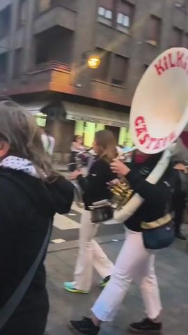 The video shows a group of people walking down a street, playing instruments. The people are mostly wearing black shirts and white pants. The street is paved with cobblestones and there are buildings on either side. There are also a few spectators watching the procession. The video appears to be taken during the daytime.