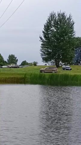 A body of water is in the foreground. On the far bank, a grassy slope leads up to trees and some buildings. Several cars are parked on the grass. In the background, there are more trees and a cloudy sky. There are a few people visible on the grass near some trees. A white van is parked near a red building with windows. A pink car is visible in front of a house with a brown roof.
