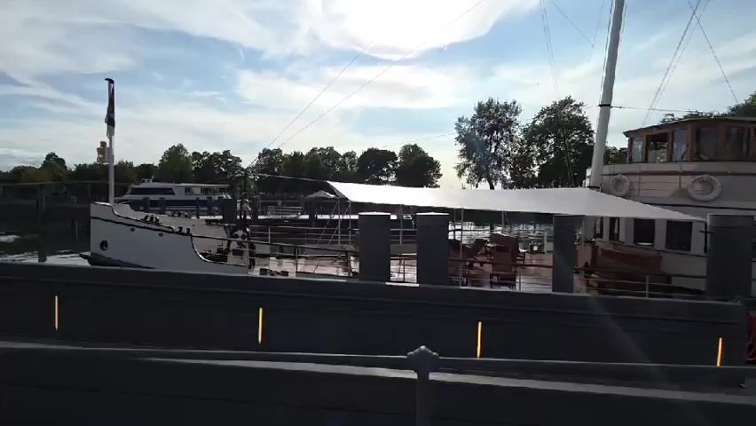 The video shows a side view of a docked steamship. The ship is white with a brown superstructure and a tall black smokestack. There is a white canopy extended over a portion of the deck. Several people are visible on the ship, including two in uniform walking on the dock. The sky is blue with some clouds, and there are trees and other boats in the background. The video appears to be taken from another dock or pier.