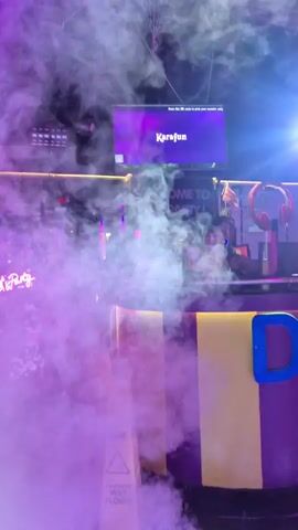 The video showcases a nightclub with a DJ booth. A DJ is visible behind the booth, equipped with DJ equipment and lights. The room is filled with fog or smoke, illuminated by colorful lights, primarily pink and purple. A screen displays the word "Karafun" along with instructions to scan a QR code. The room is decorated with various colorful elements, including what appears to be a "Big Party" sign. A person with a drink walks in front of the DJ booth through the fog. The overall atmosphere suggests a lively party scene.