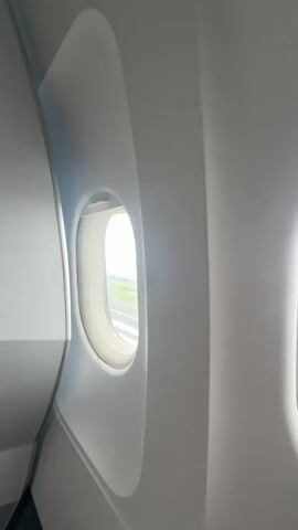 The video shows an airplane window from inside the cabin. Through the window, we see a runway and some green space under a bright sky. The plane appears to be stationary.