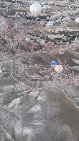 A high-angle aerial shot captures a picturesque landscape dotted with hot air balloons. The terrain below is characterized by dramatic rock formations and valleys, appearing arid and rugged. Several hot air balloons, varying in color and pattern, are scattered across the sky, some floating higher than others. The balloons are brightly colored, with notable ones including a large white balloon, a blue and white striped balloon, and a yellow and white striped balloon. The scene suggests a popular tourist destination known for hot air balloon rides, likely at sunrise or sunset given the soft lighting. The camera appears to be mounted on another hot air balloon, providing a dynamic perspective of the flight.