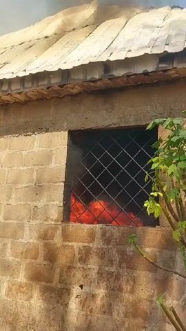 A low-angle shot shows a single-story brick building with a corrugated metal roof on fire. Black smoke billows from the roof. Flames are visible through the bars of a window. A second window has a partially open wooden shutter. A small tree is visible near the first window. The sky is visible and appears blue.