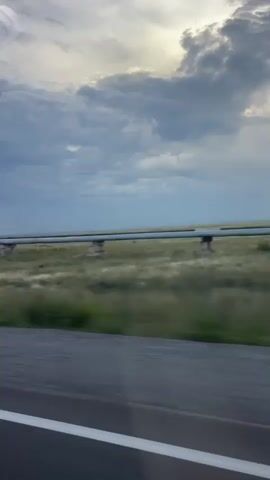 The video shows a view from a moving vehicle. A long pipe supported by concrete pillars runs horizontally across the frame in the middle distance. The pipe is in a grassy field under a cloudy sky. The foreground consists of a paved road with a white line. A white vehicle with large tires passes by on the road. The time of day appears to be dusk or dawn, as indicated by the lighting and clouds.