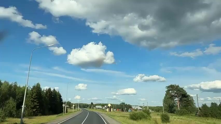 A car is driving down a two-lane road. The sky is blue with white and gray clouds. There are trees and houses on both sides of the road. There are streetlights along the road.