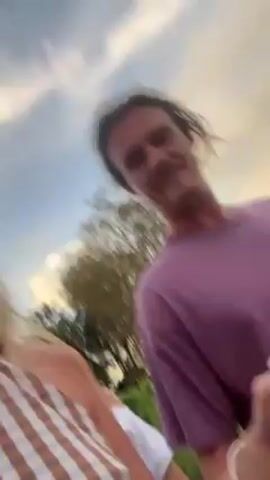 Three individuals are captured in a brief, shaky video clip. The person holding the camera is a blonde woman, wearing a brown and white striped top. To her right, a man wearing a purple t-shirt gives the peace sign with his left hand, before dropping it. Behind them is another young woman wearing a white top, only partially visible. They are outside, with trees and a partly cloudy sky visible in the background. The video appears to be a casual greeting to the camera.
