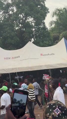 The video shows a crowded outdoor event. Many people stand beneath a white and blue tent bearing the name VEN-BAWA. A person in a colorful traditional costume with a mask walks among the crowd. Some people are drinking water. Trees and buildings surround the event. The atmosphere seems festive and lively.