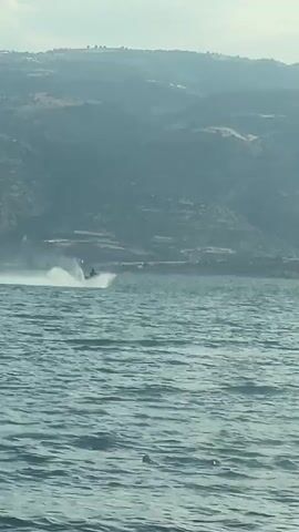 A person on a jet ski is riding across a body of water. The jet ski is creating a large spray of water behind it. In the background, there are mountains. The sky is overcast.