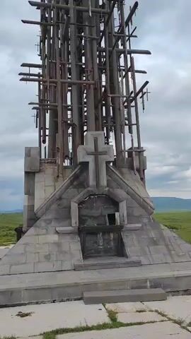 The video shows a tall, unfinished cross structure made of metal beams and wooden planks. The cross is located in an open, grassy field under a cloudy sky. The base of the cross is a stone structure with a carved cross and an opening. The camera pans up the cross, revealing its intricate and somewhat rustic construction.