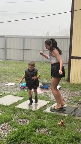 A young child wearing a black puffer vest and black boots, and a woman wearing a white tank top and black shorts are dancing outside in the rain. The child is on a stone path, and the woman is standing next to him on the grass. There is a metal fence and a building in the background. The ground is wet from the rain, and there are some toys scattered on the grass.