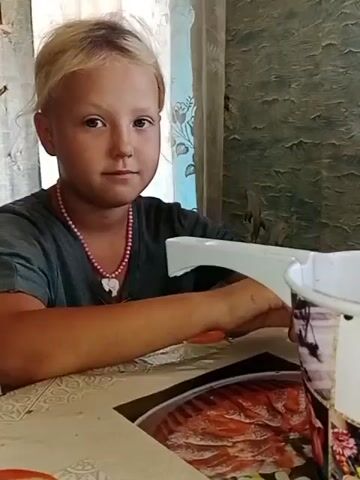 A young girl with blonde hair and a pink necklace sits at a table. She is wearing a dark t-shirt. Her right hand rests on the table. A white object is partially visible in front of her. A bucket with an image on it is to her right. The background consists of a patterned wall and a curtain. The girl smiles at the camera.