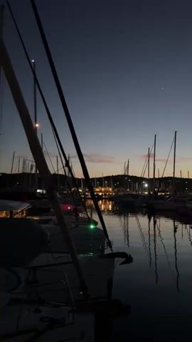 A static, vertical shot captures a marina scene at dusk. Numerous sailboats, primarily visible as dark silhouettes of their masts and hulls, are docked in still water. The masts are reflected clearly on the water's surface. In the foreground, part of a boat's bow with a green light is visible on the left. Across the water, lights from distant buildings or structures on the shore illuminate the background. The sky transitions from a deep blue overhead to a lighter, orangey-pink hue near the horizon, with two faint stars visible in the upper right.