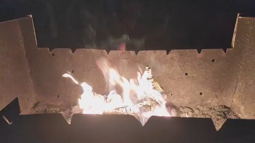 A close-up, low-angle shot captures a crackling campfire in a metal brazier. The flames are a vibrant mix of white, yellow, and orange, licking upwards and illuminating the surrounding logs. The logs themselves are charred and fragmented, showing the intense heat of the fire. The brazier appears to be made of rough, dark metal with a serrated top edge, and the background is dark, suggesting it is nighttime. The scene conveys a sense of warmth and perhaps a cozy outdoor gathering. There are no people visible in the frame. The location context is Kolpakovo, Russia.