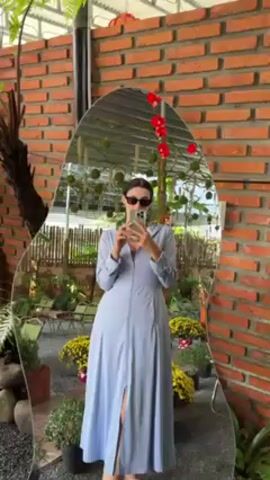 The video shows a person taking a selfie in a uniquely shaped mirror. The person is wearing a light blue, long-sleeved dress and sandals. They are holding a smartphone and looking at the screen. The mirror reflects a garden setting with plants, flowers, and outdoor furniture. Behind the mirror is a brick wall. The camera then pans to show more of the garden area, including tables and chairs under a covered patio. The environment appears to be a cafe or outdoor dining area.
