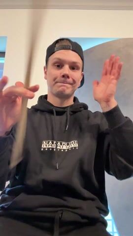 A young man wearing a black baseball cap and black hoodie juggles a drumstick. He holds the drumstick vertically in front of him, then holds it horizontally, then waves it in the air above his head with a sudden yell. The man is indoors and is facing the camera. The background consists of white walls and gray cushions.