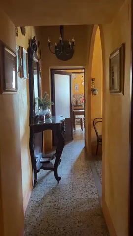 The video shows a room in a house. The camera moves from a hallway, which is decorated with framed pictures and a dark wooden table, into a living room. The walls of the living room are a yellow-orange color. A brown sofa sits in the middle of the room. There's a dark wooden cabinet with a lamp and other decorative items. A TV is visible near the window. The ceiling is wooden with exposed beams. Decorations like plates are visible on the wall. The hallway leads to another room, visible through an open door.