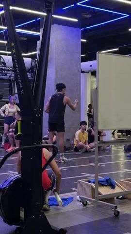 A group of people are in a brightly lit indoor gym. A man wearing a black tank top and black shorts stands center frame, facing away from the camera, and appears to be instructing a class. To his left, a woman with purple hair wears a white jacket and blue shorts, and a woman in a red tank top is partially visible in the foreground, sitting on a rowing machine. Several other individuals are seated on the floor in the background, facing the instructor. A whiteboard stands to the right of the instructor. The environment includes gym equipment such as rowing machines and various exercise apparatuses. The lighting is a mix of natural light and artificial blue and white neon lights.