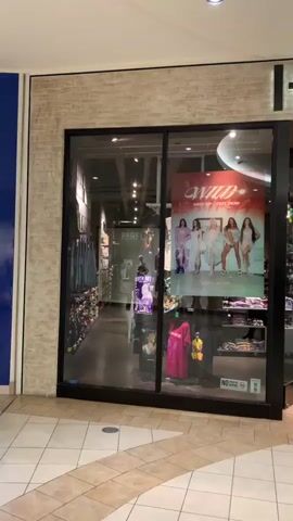 The video shows the exterior of a Hot Topic store in a mall. The store has large glass windows displaying merchandise. Inside, racks of clothing and shelves stocked with various items are visible. A large poster featuring women in swimsuits is displayed in the left window, with the word "WILD" in large red letters at the top. In the right window, a circular sign advertises a "Funko" promotion offering 30% off. The store's name, "HOT TOPIC," is prominently displayed in large, illuminated letters above the entrance. The floor in front of the store is tiled with a checkerboard pattern.