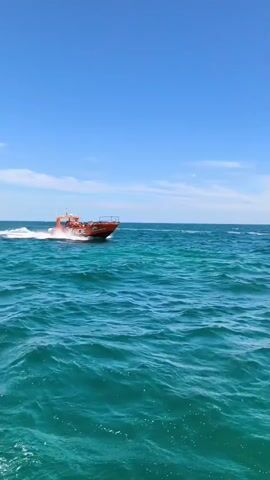 The video shows a bright orange boat with people on board speeding through turquoise ocean waters under a clear blue sky. The boat appears to be a tourist vessel, possibly a jet boat or a similar high-speed craft designed for exhilarating rides. The water is choppy, creating white wakes behind the boat, indicating it's moving at a considerable speed. Several passengers are visible, some appearing to be holding on as they ride. The overall scene suggests a sunny day, likely mid-day given the brightness and clear sky. The ocean's color suggests a tropical or sub-tropical location with clear, shallow waters. Text is visible on the side of the boat, reading "X RIDE" and potentially other words in a smaller font. The dynamic movement of the boat and the open water setting evoke a sense of adventure and fun. The location context provided, Lagoa, Portugal, aligns with the visual cues of coastal waters and a sunny climate.