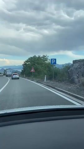 The video shows a view from inside a car driving on a paved road. The road curves to the right, and there are white dashed lines marking the lanes. On the right side of the road, there is a metal guardrail, trees, and a sign indicating a curve. In the distance, mountains and a body of water can be seen under a cloudy sky. Several cars are driving on the road ahead, including a white truck and a white car. The driver's dashboard is visible in the foreground.