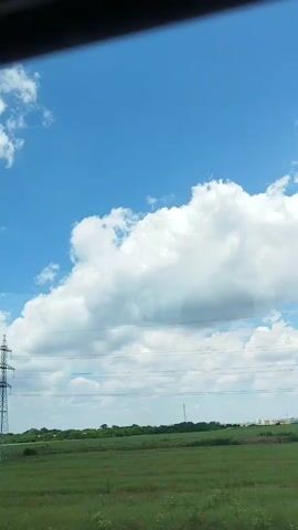 The video shows a view from a moving vehicle, likely a car or train, looking out at a rural landscape. The sky is bright blue with fluffy white clouds. A tall, metallic power line tower is visible on the left side of the frame. In the middle ground, there is a green field with scattered red flowers, possibly poppies. A road sign is briefly visible on the right side, with the text "S.O.S. 112" on it. The scene suggests a daytime journey through the countryside under a clear sky.