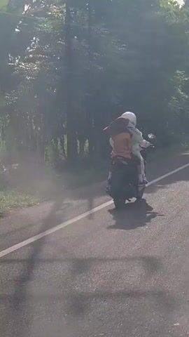 The video shows two people riding a motorcycle on a paved road. The person in the front is wearing a white helmet and a light-colored jacket. The person in the back is wearing an orange shirt and a helmet. The motorcycle is moving away from the camera, and there is dust or smoke trailing behind it. The environment appears to be a rural area with trees and vegetation on the side of the road. Power lines are also visible in the background. The road has a white line marking the edge.