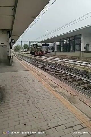 The video captures a view from a train station platform looking down the tracks. In the foreground, a brick-paved platform with a yellow safety line is visible. Several sets of train tracks run parallel to the platform. In the mid-ground, a train is parked on one of the tracks. The train appears to be a locomotive with some carriages or a cargo load behind it. The station building is visible in the background, with a roof and windows. The environment appears overcast or hazy, suggesting cloudy weather. The timestamp on the video indicates it was recorded on July 2, 2026, at 09:09. There are no people clearly visible interacting with the scene in the provided clip.