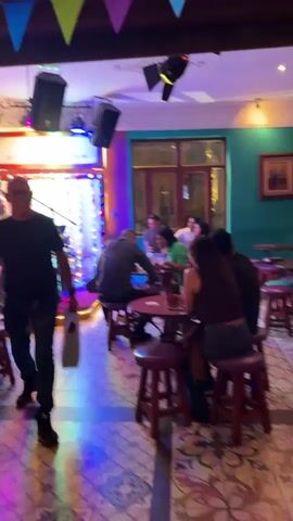 The video takes place inside a cozy bar with a live band. A man in a black shirt, glasses, and dark pants walks through the bar carrying a tan shopping bag, heading towards a stage where a band is setting up.  Several tables and chairs are arranged around the room, occupied by small groups of people enjoying drinks and conversation.  The bar is adorned with colorful flags and string lights, creating a festive atmosphere.  As the camera moves, a smiling man wearing glasses, a black shirt, and a beard faces the camera directly, briefly filling the frame.  The overall environment is vibrant and social, suggesting a relaxed evening at a local establishment.