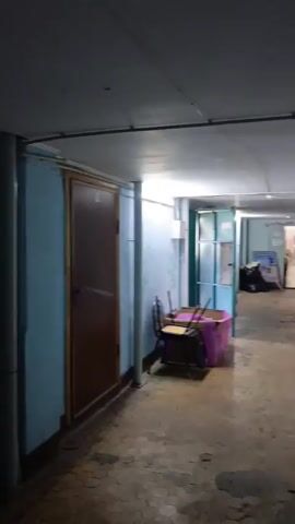 The video shows a hallway with blue walls. A blue door is visible on the left. Further down the hallway, a red fire extinguisher hangs on the wall. There is another door on the right side of the hallway, which is dark inside. The hallway appears to be indoors and possibly in a building. The camera is moving, suggesting someone is walking through the hallway. The lighting is dim in some areas. There are pipes and conduits running along the walls and ceiling. There are no people visible in the video, and no actions or interactions are depicted.