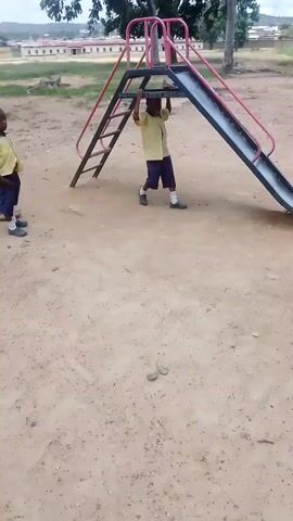 Three children are playing on a playground with a slide. One child in an orange shirt is standing at the bottom of the slide. Another child in a yellow shirt is on the slide, upside down, hanging by their hands. A third child, also in a yellow shirt, is standing to the side. The ground is sandy and dusty. In the background, there are buildings and trees.