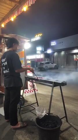 A young person wearing a black t-shirt and black pants is standing on the right side of the frame, facing a grill with meat skewers on it. They are holding a stick with tongs and appear to be cooking the meat over hot coals. Smoke rises from the grill. In the background, there is a white car, buildings with illuminated signs, and string lights overhead. The scene is dimly lit, suggesting it is nighttime. There is a black bucket filled with charcoal near the grill.