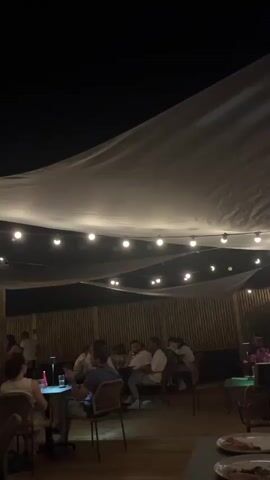 The video shows an outdoor seating area at night. There are several groups of people sitting at tables. A white fabric canopy is stretched overhead, supported by wooden poles, and illuminated by a string of lights. In the background, there is a bamboo fence and a building with windows. Some people are standing and conversing, while others are seated and eating or drinking. The overall atmosphere appears to be relaxed and social.