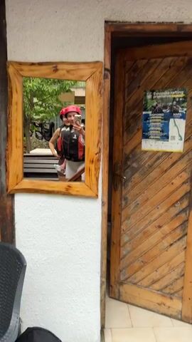 Two women are taking a selfie in a mirror. They are both wearing life vests and helmets, suggesting they are about to participate in an outdoor activity. The woman in the foreground is holding a phone and making a playful face. The woman behind her is also smiling. The mirror is framed with rough-hewn wood, and the reflection shows trees and foliage in the background. To the right of the mirror is a wooden door with a sign on it.
